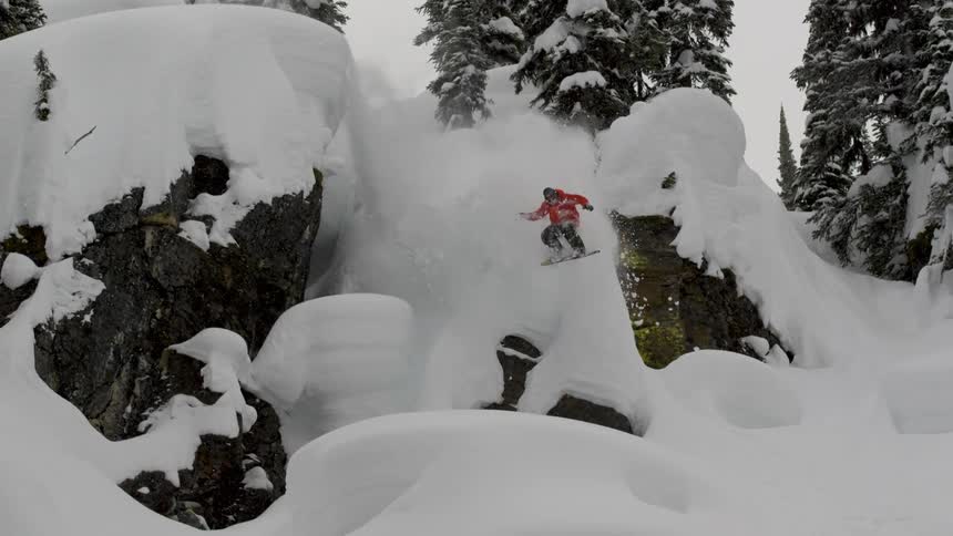 A snowboarder jumps of a cliff into pillows of snow
