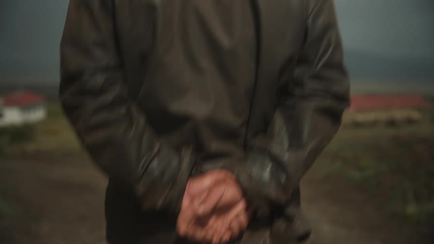 Man with a hat and leather jacket walking backwards in a rural area