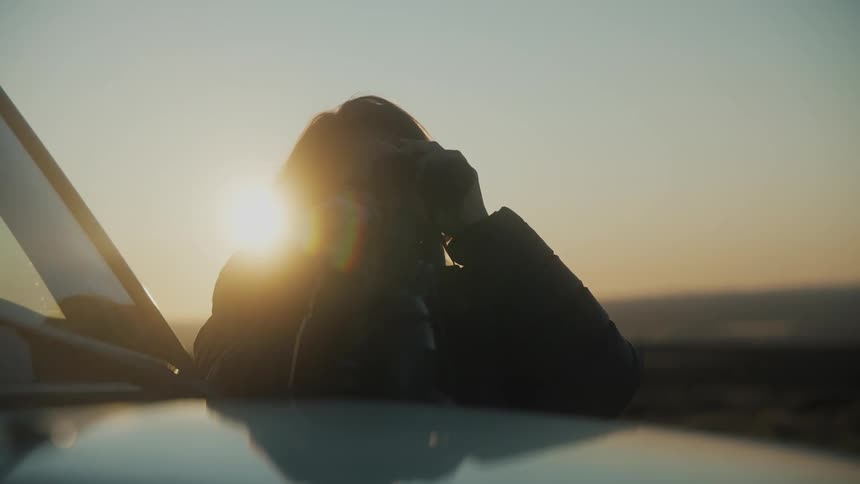 A woman leaning on a car observing the landscape with binoculars