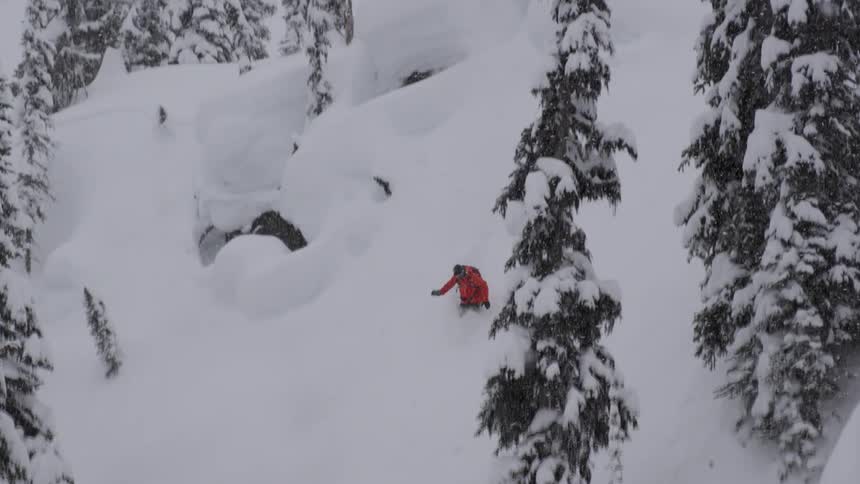 A snowboarder jumps into powder in steep trees