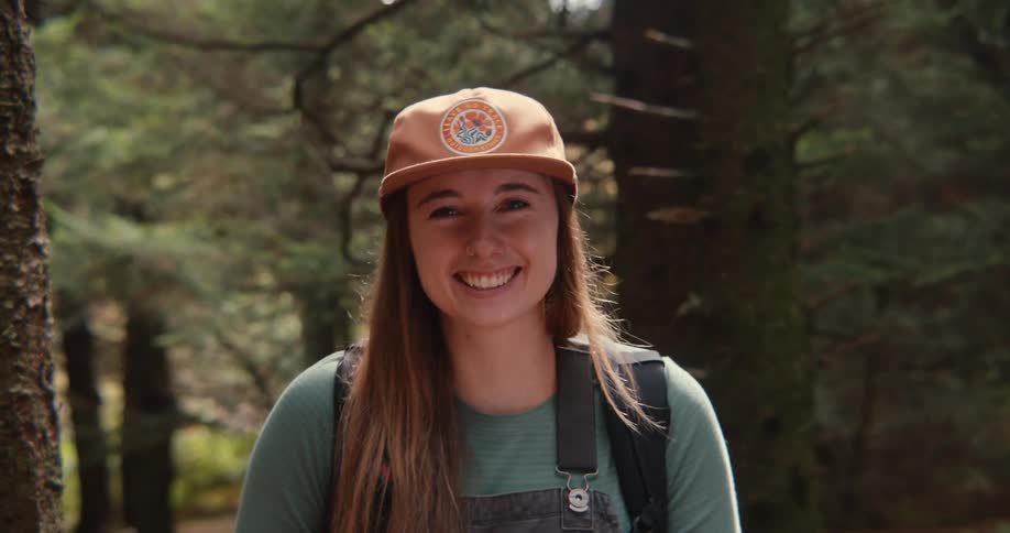 Smiling white woman with a cap and backpack amidst the trees in woodland