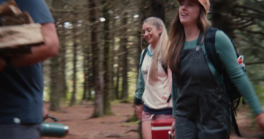 Friends smile while carrying coolers and firewood through a conifer forest