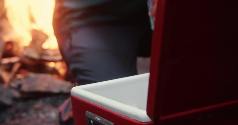 Man crouching down and grabbing beverage cans from a cooler next to a campfire in the nature