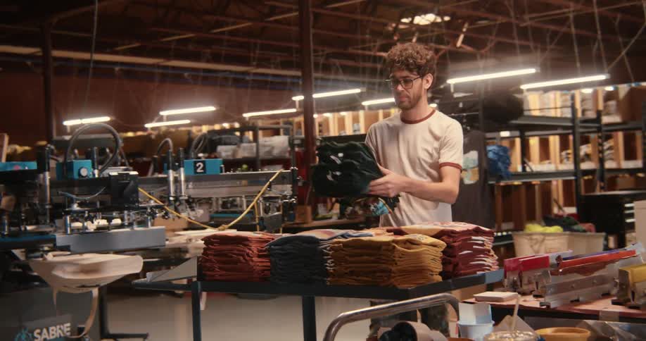 Casual dressed employee in a screen printing shop carries a stack of shirts to a cart with other color sorted stacks of shirts surrounded by machines and sets them down