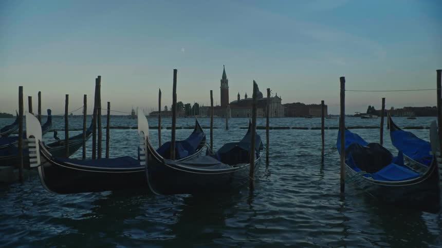 Gondolas bob in water as boats pass historic buildings in the background at dusk