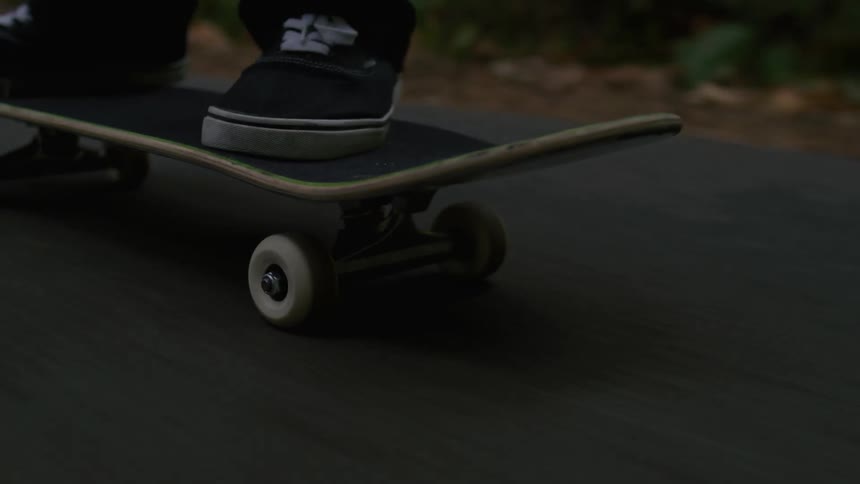 Black converse style shoes on a skateboarder riding on a skateboard down a paved road in a wooded area right before dark
