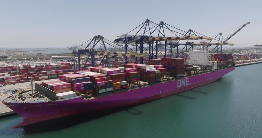 An aerial view of a large cargo ship with stacked trailers in a large harbor region of a city