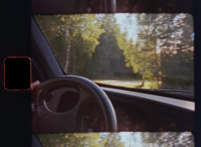 Hands of driver driving a car turning the steering wheel to leave the main road and make a turn onto a small gravel road in a forest as he arrives or leaves on a sunny day with bright flickering light and lens flare on vintage Super 8mm film