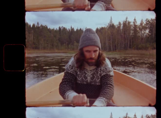 Frontal portrait of a man sitting in an old orange rowing boat and paddling with wooden oars on a forest lake through a bay with water lilies towards the shore with trees on a sunny autumn day on vintage film