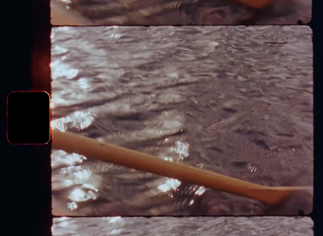 Portrait of a smiling man with short beard and long brown hair wearing a beanie hat and a woolen pullover or sweater paddling in a canoe or boat on a river or lake on a sunny day and the rippling surface of the water reflects sunshine