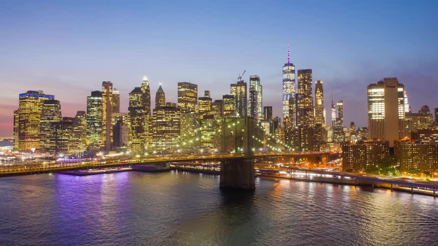 Bridge over a bay connects to the New York skyline lit up under a purple sky at dusk with blurred moving lights of traffic speeding along the roads