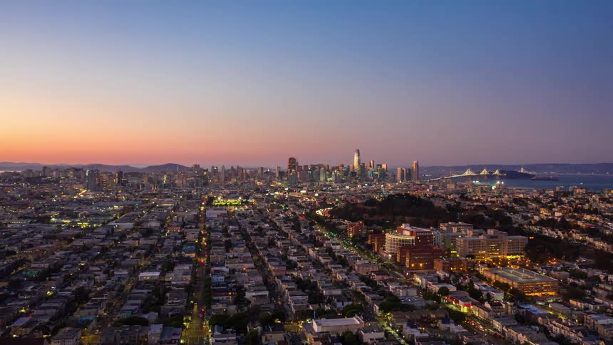 Low aerial over a large city by the ocean with blurred lights from quick moving traffic from dawn to dusk against a blue purple and gold sky