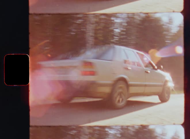 Passing vintage car or old white vehicle drives down the small gravel road by the forest as the sun goes down in the evening during sunset on a warm summer day at the end of a carefree road trip on the rural countryside on vintage S8 film