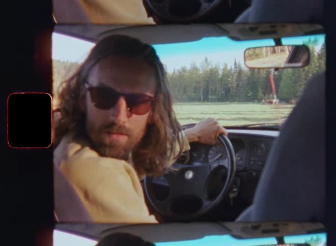 A brunette man with long hair and sunglasses sitting in a retro vehicle looks over his shoulder and backs up or reverses on a country road in a rural area as the bright flickering sun shines onto his face on vintage Super 8mm film
