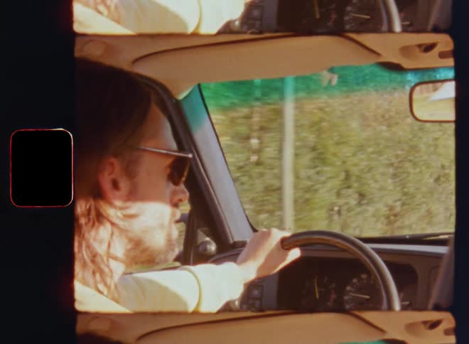 A brown-haired man with glasses is driving a car on a sunny day looking around and passing near trees next to the street while enjoying the road trip and careless happy summer vibes on vintage Super 8 mm film