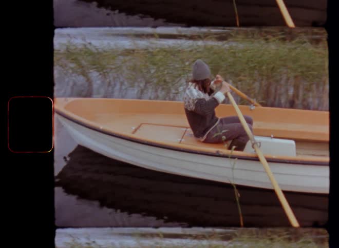 Young brunette man paddling an orange and white rowing boat with pair of paddles or oars through the reeds or cane growing at the shore or edge of forest lake or river enjoying leisure time in the outdoors