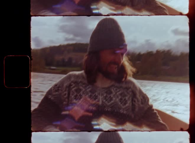 A happy man with long hair and beanie hat paddling a canoe or rowing boat on a lake during a sunny day while enjoying the outdoor activity and laughing on vintage Super 8 film with retro summer or autumn vibes