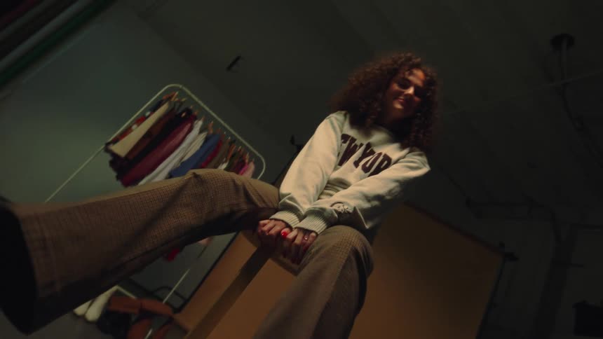 A young woman sits and moves her legs and behind her there are several clothes on the hanger
