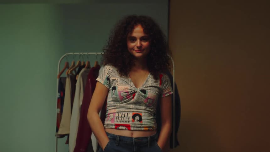 A young woman with curly hair models a shirt while standing indoors