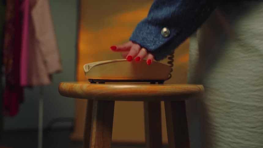 Basic landline phone on a stool and is answered by a woman with red nail polish who picks up the phone and walks away while light from a setting sun cast shadows on the wall