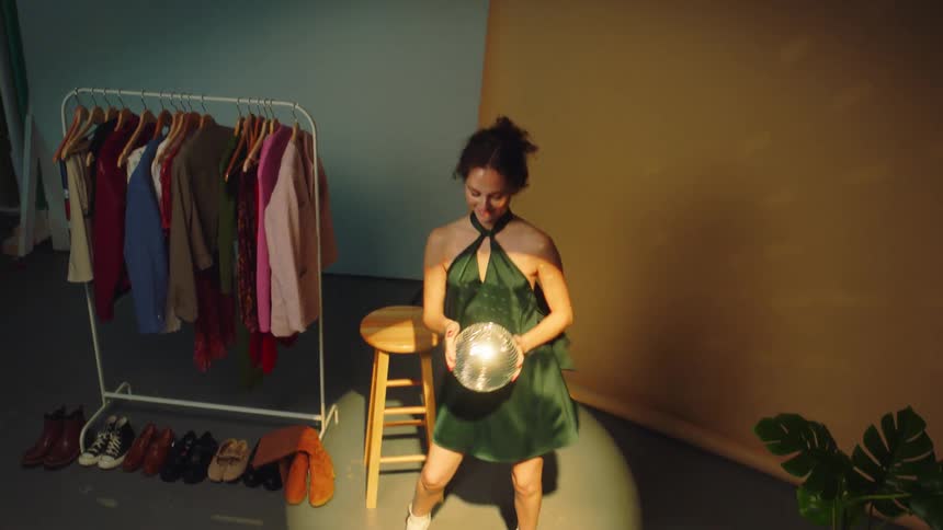 Woman dancing in spotlight near rack of clothes holds a small mirrorball