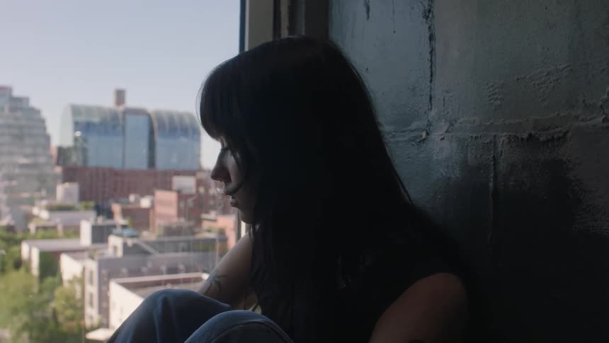 Young woman sitting on the floor of a building looking out the window at a city looking disheartened