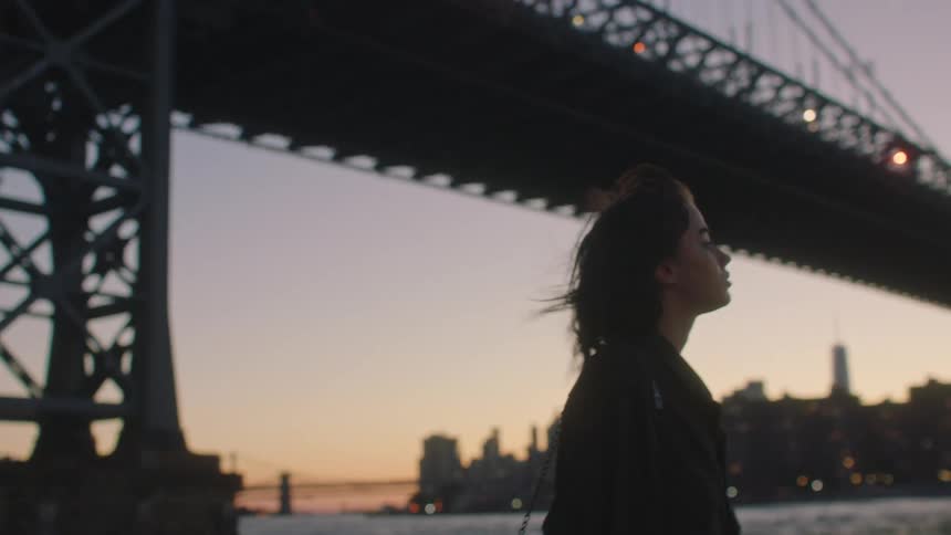 Young woman standing next to a river with a bridge behind her in a metropolitan area during dusk or dawn