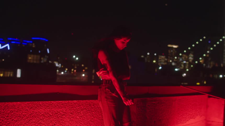 A young woman stands on a city roof at night head bowed fidgeting restlessly