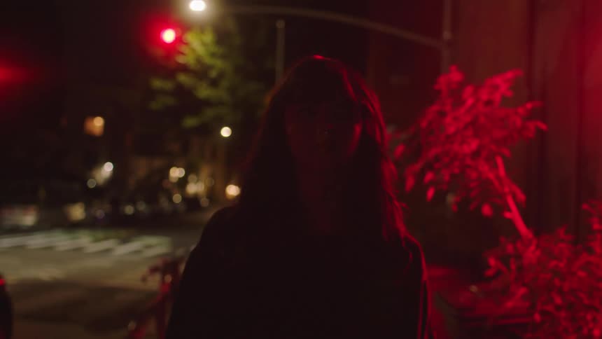 Brunette haired young woman with a frown walks down a city street past red neon sign at night