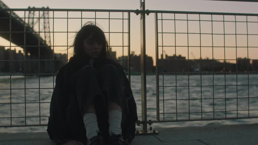 A young woman walks to the fence next to a river and sits down against it