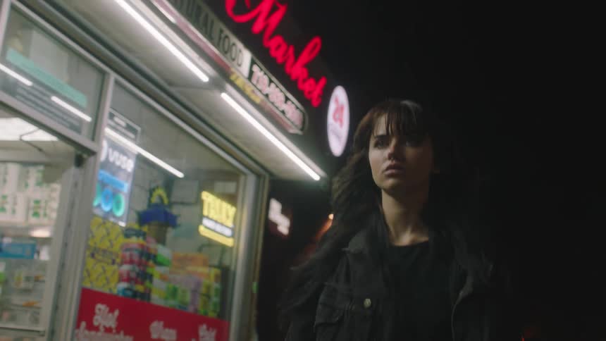 Young brunette adult female walks down a city block past a well lit convenience store at night
