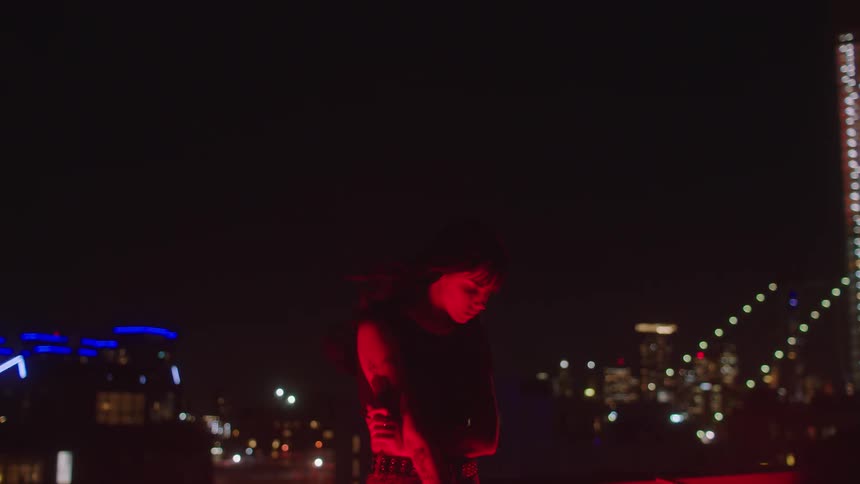 Young woman with one arm crossed looking down with the wind blowing on her face in an outdoor setting in a metropolis at night