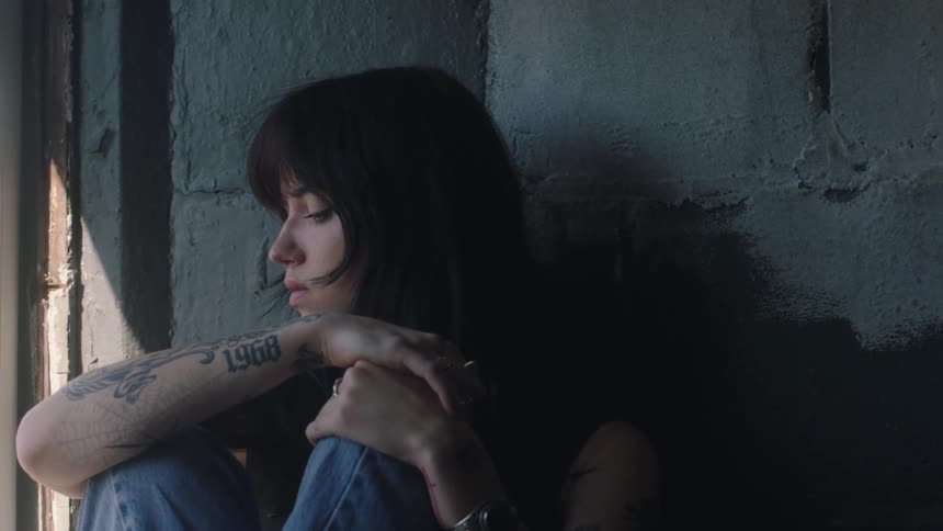 A young tattooed woman is sitting and leaning against a wall with a sad face