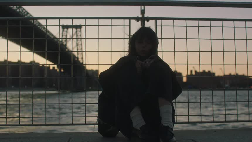 Teenage girl walks to fence and sits down facing away from the river and bridge