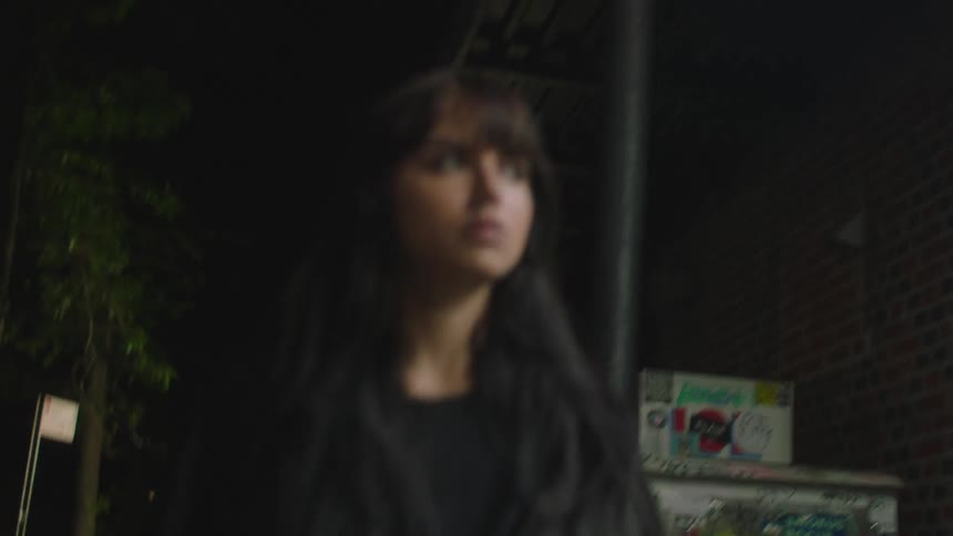 Woman with dark hair and jacket walks down the street at night