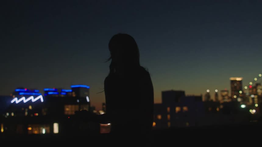 A woman smoking a cigarette on top of a building in the city at night