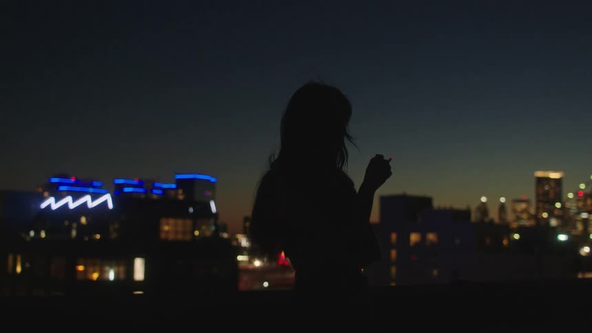 Silhouette of a woman smoking in front of the city at night