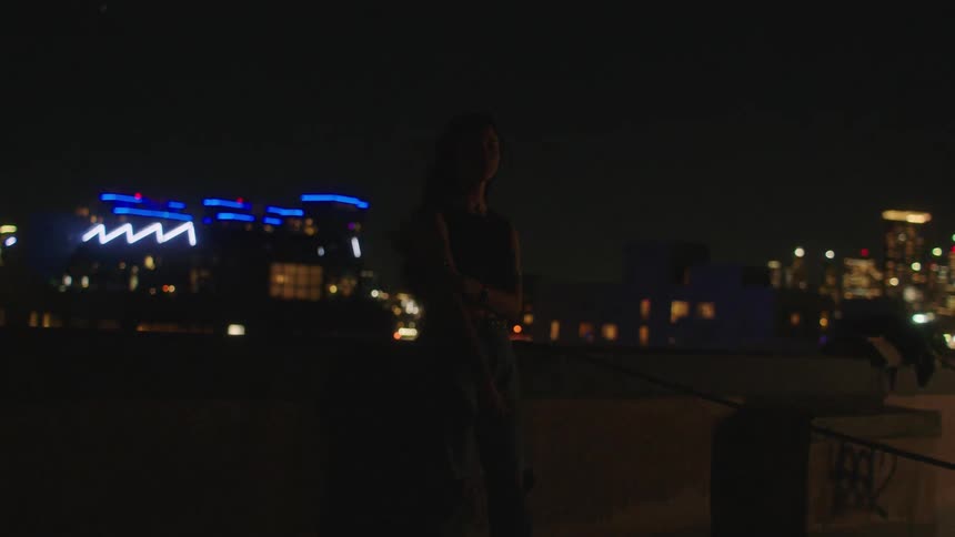 Woman leaning against concrete in the middle of a city at night with lights reflecting beneath her