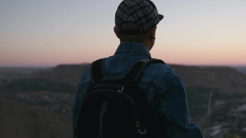 A man stands on a hillside at dawn and adjusts his cap