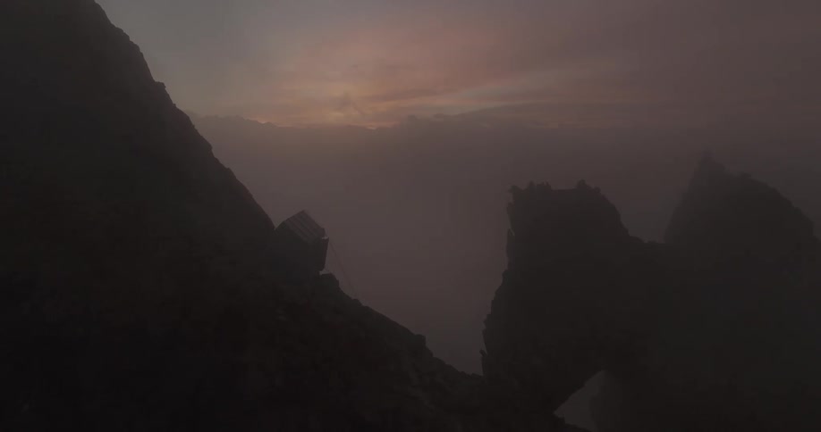 A rocky formation on a cliff with a house on the side with fog and a sun hiding behind a mountain range in the distance