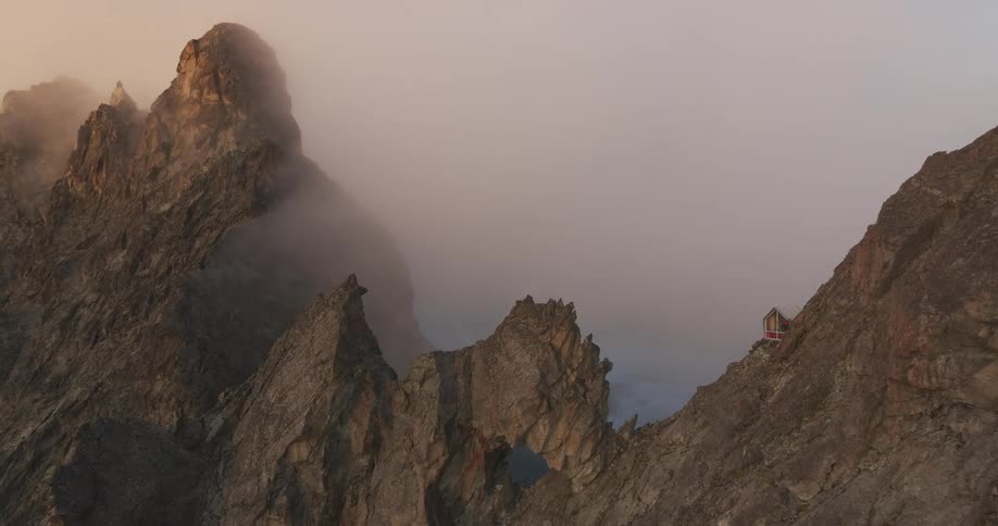 Tent balances on steep mountain slope over rock formation containing a hole under red tinted low lying clouds