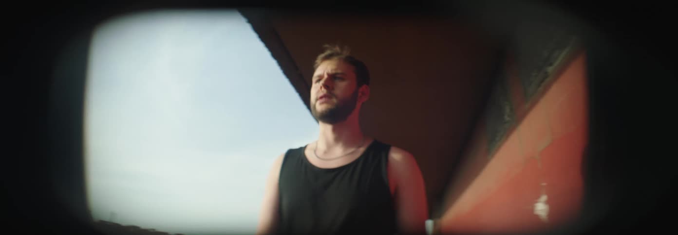 Bearded man with a necklace and a black tank top walks by the window shaking his head