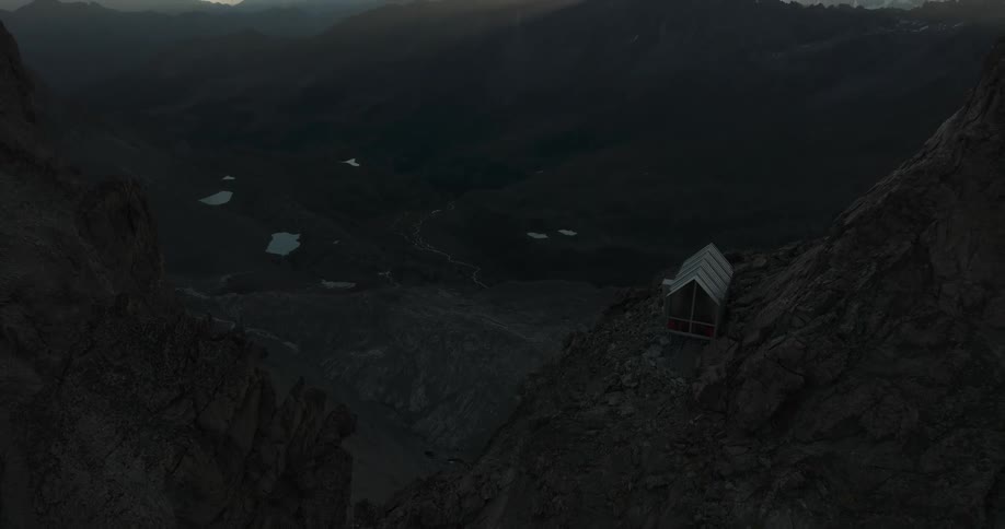 Aerial moving out away from single house on the edge of a mountain cliff with mountains on the horizon under a bright sky with the sunrising behind a dark cloud