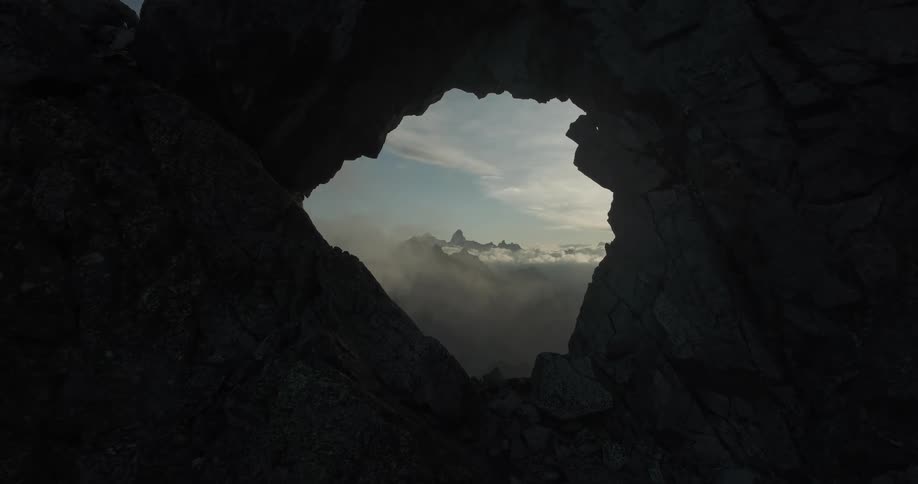 Through a natural hole in a rock formation with clouds on the other side darking as the sun is setting