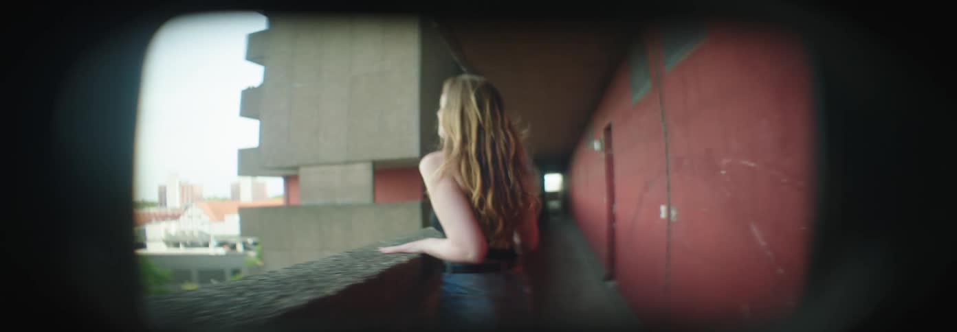 Blonde woman with her back walking down a corridor towards a balcony and talking to someone outside