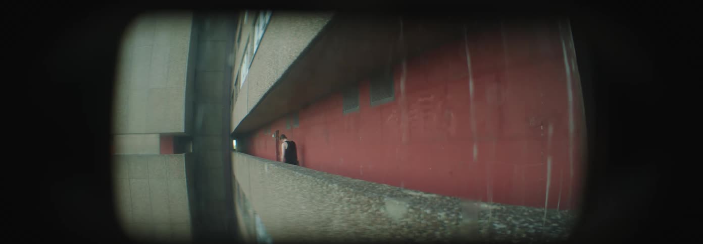 Man wearing tank top walks along outdoor hallway leans against wall and crouches down