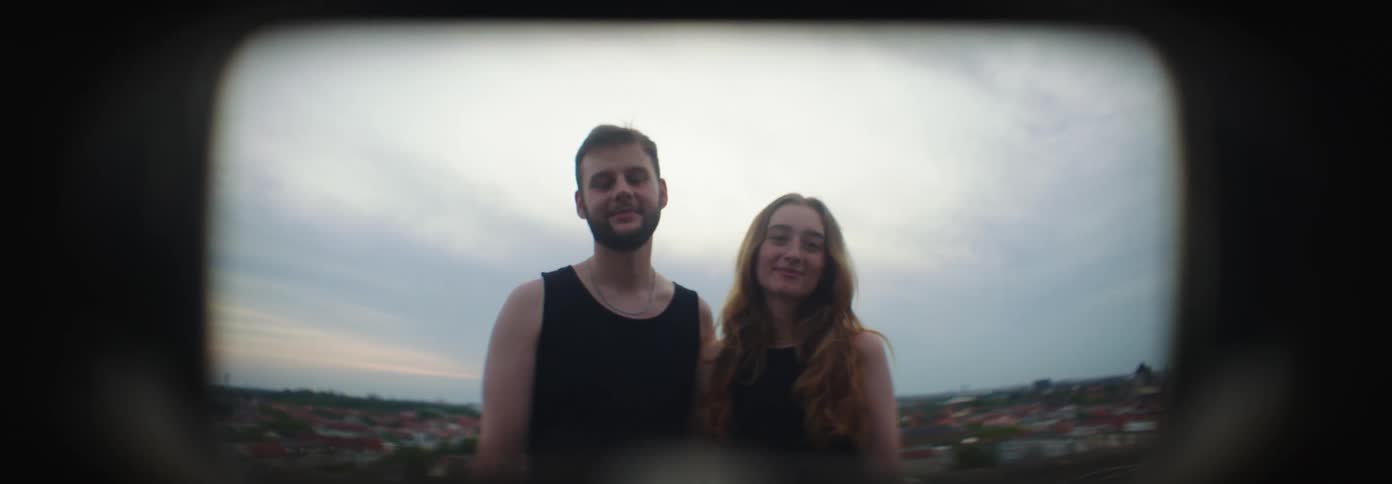 Couple side by side embracing and smiling with a suburban cityscape in the background under a cloudy sky