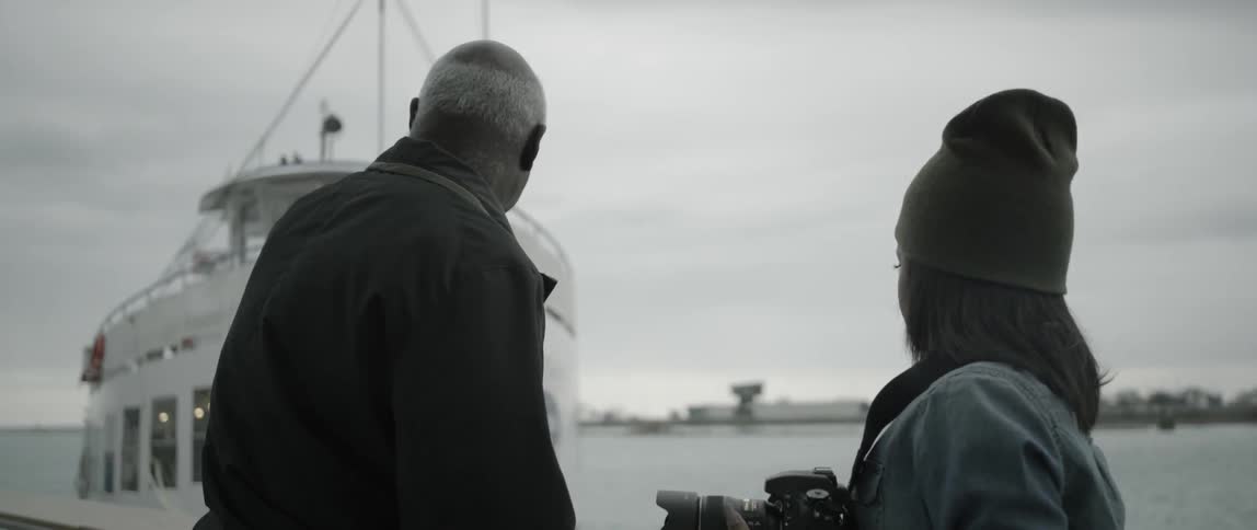 A woman holds a camera near a man as a boat gets near the pier