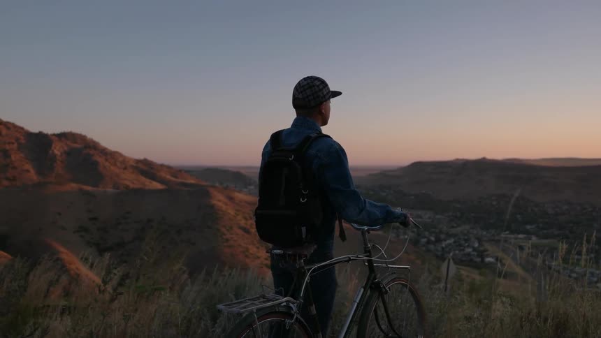 A man stands with his bike on a hill top at dawn