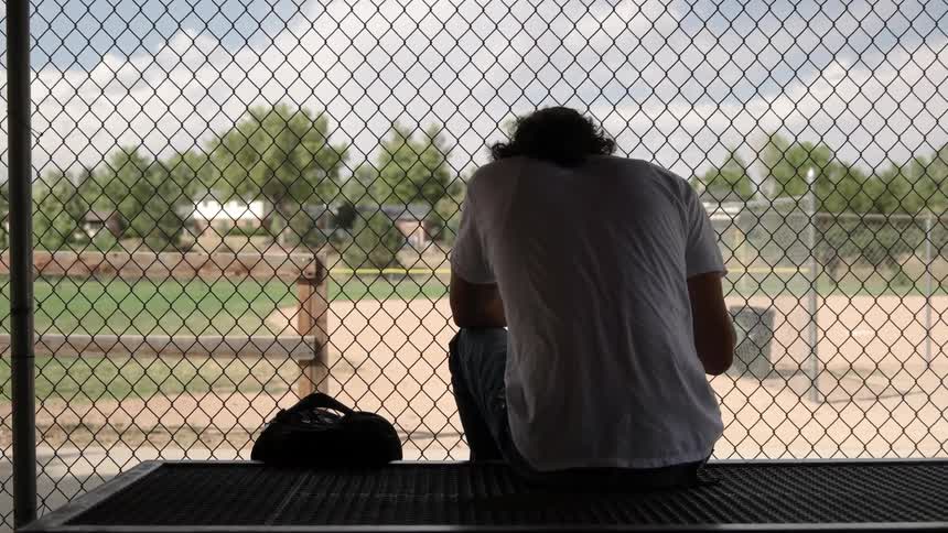 Man sits on a picnic table outside near a chain link fence